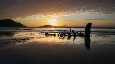 Swansea U 'daki Gower yarımadasındaki Worms Head' in önünde, Rhossili körfezindeki Helvetia 'nın enkazının üzerinde gün batımı.