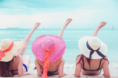 Seksi bikini vücudu. Üç Asyalı kadın kumsalda uzanmış şapkalarla denizin tadını çıkarıyorlar..