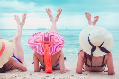 Seksi bikini vücudu. Üç Asyalı kadın kumsalda uzanmış şapkalarla denizin tadını çıkarıyorlar..