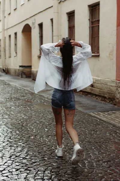 Yağmurlu bir havada sokakta yürüyen bir kadın. Beyaz gömlekli ve kot şortlu ıslak bir kadın. Yağmurlu bir şehirde esmer bir kız.