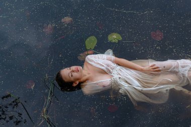 Suyun Huzurlu Vücudu pozunda, Beyaz Elbiseli Serene Kadını. Ormanda uzun ve şeffaf bir gelinlik giymiş kadın ya da gelin. Göl, gölet ya da deniz yakınlarındaki vahşi sahil. Su zambakları. Güzel kız..