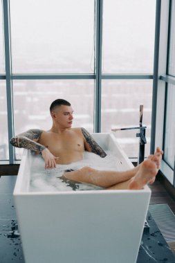 Küvette rahatlayan bir adam. Şehir manzaralı panoramik pencere, banyo içi lüks. Köpüklü dövmeli adam..