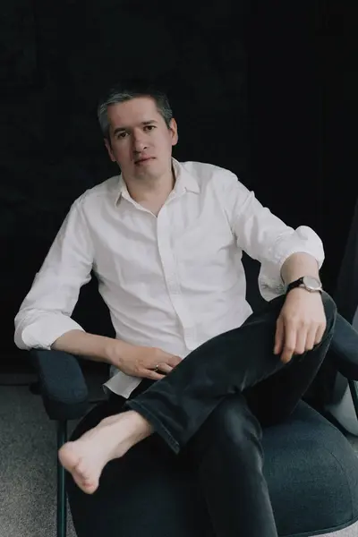 Sandalyede oturan bir adam. Evin içinde lüks bir yer. Beyaz gömlekli ve siyah kotlu bir erkek..