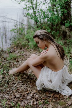 Serene Woman in a White Dress by a Tranquil Body of Water oturur ve poz verir. Ormanda uzun ve şeffaf bir gelinlik giymiş kadın ya da gelin. Laker, gölet, deniz yakınlarındaki vahşi sahil. Yaz titreşimleri. Güzel kız.
