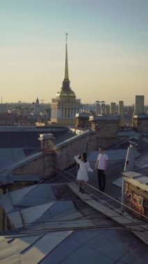 Bir adam ve bir kadın çatıda yürüyor, sarılıyor ve öpüşüyorlar. Saint-Petersburg, Rusya 'da çatı. Deniz Kuvvetleri 'ne bakın. Gün batımında şehirde romantik bir çift buluşması. Dikey