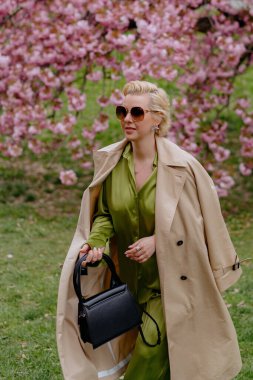 Çiçek açan pembe kiraz çiçeklerinin önünde, yeşil saten elbiseli şık sarışın bir kadın zarafet ve zarafet yayar, baharın ve doğanın güzelliğini büyüleyici bir şekilde yakalar.