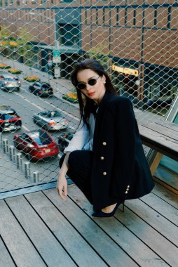 Siyah ceket ve güneş gözlüğü takmış şık bir kadın şehrin arka planına karşı şehir balkonunda kendine güvenen bir duruş sergiliyor şık ve sofistike bir görünüm sergiliyor.