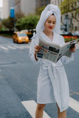 Rahat bir bornoz ve havlu giymiş şık bir kadın, canlı bir şehir ortamında gazetesini okumaktan zevk alıyor. New York 'ta şehir caddesi.