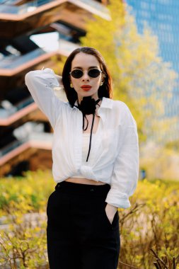 Beyaz bluz ve siyah pantolon giyen kendine güvenen bir kadın modern bir şehir geçmişine karşı poz veriyor. Gelişmişlik ve zarafeti somutlaştırıyor. Çağdaş ortamda kendine güven ve tarz yayıyor.
