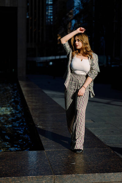 A stylish woman entrepreneur, elegantly dressed, confidently walks near a modern office building, embodying empowerment, determination, and style under the sun in the bustling city