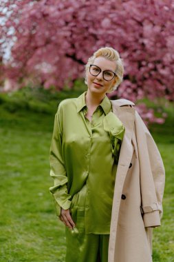 Çiçek açan pembe kiraz çiçeklerinin önünde, yeşil saten elbiseli şık sarışın bir kadın zarafet ve zarafet yayar, baharın ve doğanın güzelliğini büyüleyici bir şekilde yakalar.