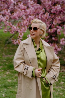 Çiçek açan pembe kiraz çiçeklerinin önünde, yeşil saten elbiseli şık sarışın bir kadın zarafet ve zarafet yayar, baharın ve doğanın güzelliğini büyüleyici bir şekilde yakalar.