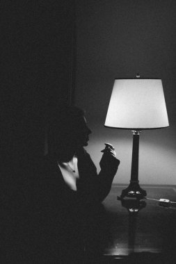 In a dimly lit room, a silhouette of a person stands by a table lamp, creating a striking contrast of light and shadow in the black and white image, evoking a sense of mystery and intimacy clipart