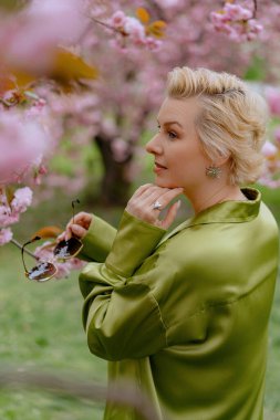 Çiçek açan pembe kiraz çiçeklerinin önünde, yeşil saten elbiseli şık sarışın bir kadın zarafet ve zarafet yayar, baharın ve doğanın güzelliğini büyüleyici bir şekilde yakalar.