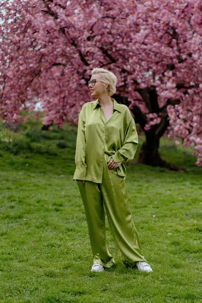 Çiçek açan pembe kiraz çiçeklerinin önünde, yeşil saten elbiseli şık sarışın bir kadın zarafet ve zarafet yayar, baharın ve doğanın güzelliğini büyüleyici bir şekilde yakalar.