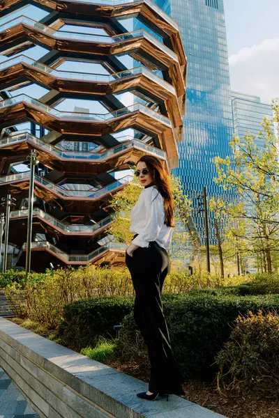 Beyaz bluz ve siyah pantolon giyen kendine güvenen bir kadın modern bir şehir geçmişine karşı poz veriyor. Gelişmişlik ve zarafeti somutlaştırıyor. Çağdaş ortamda kendine güven ve tarz yayıyor.