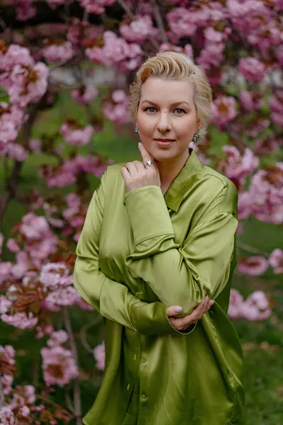 Çiçek açan pembe kiraz çiçeklerinin önünde, yeşil saten elbiseli şık sarışın bir kadın zarafet ve zarafet yayar, baharın ve doğanın güzelliğini büyüleyici bir şekilde yakalar.