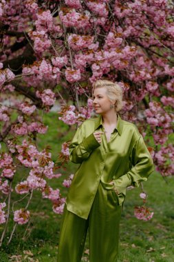 Çiçek açan pembe kiraz çiçeklerinin önünde, yeşil saten elbiseli şık sarışın bir kadın zarafet ve zarafet yayar, baharın ve doğanın güzelliğini büyüleyici bir şekilde yakalar.