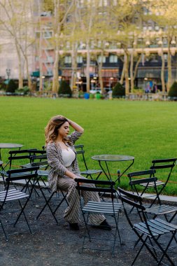 Şık bir kadın şehir parkındaki şık bir teras masasında, gökdelenlerin ve yeşilliklerin ortasında sakin ve sofistike bir rahatlama sahnesi yaratarak eğleniyor.