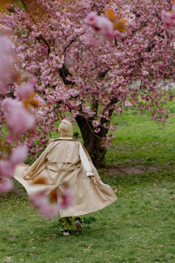 Çiçek açan pembe kiraz çiçeklerinin önünde, yeşil saten elbiseli şık sarışın bir kadın zarafet ve zarafet yayar, baharın ve doğanın güzelliğini büyüleyici bir şekilde yakalar.
