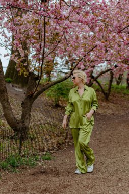 Çiçek açan pembe kiraz çiçeklerinin önünde, yeşil saten elbiseli şık sarışın bir kadın zarafet ve zarafet yayar, baharın ve doğanın güzelliğini büyüleyici bir şekilde yakalar.