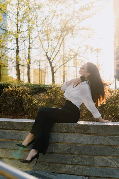 Beyaz bluz ve siyah pantolon giyen kendine güvenen bir kadın modern bir şehir geçmişine karşı poz veriyor. Gelişmişlik ve zarafeti somutlaştırıyor. Çağdaş ortamda kendine güven ve tarz yayıyor.