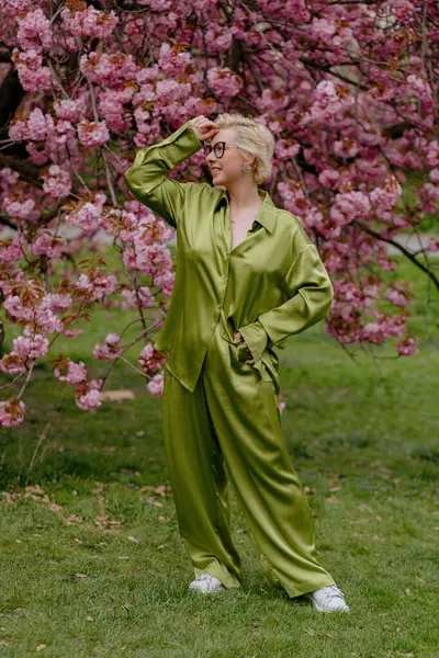 Çiçek açan pembe kiraz çiçeklerinin önünde, yeşil saten elbiseli şık sarışın bir kadın zarafet ve zarafet yayar, baharın ve doğanın güzelliğini büyüleyici bir şekilde yakalar.