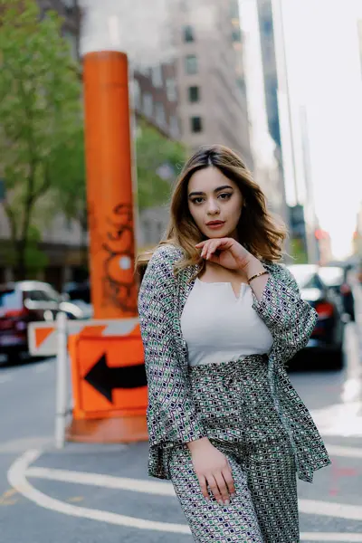Şık bir kadın şehir caddesinde güven içinde poz veriyor. Şık kıyafetler giyiyor ve içinde binalar ve buharlı havalandırma bulunan bir şehir arka planının önünde duruyor. Mükemmel bir şehir modası fotoğrafı..