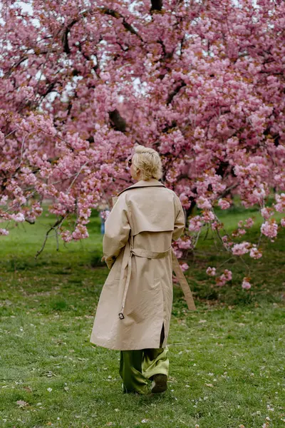 Çiçek açan pembe kiraz çiçeklerinin önünde, yeşil saten elbiseli şık sarışın bir kadın zarafet ve zarafet yayar, baharın ve doğanın güzelliğini büyüleyici bir şekilde yakalar.