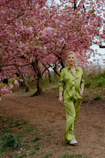 Çiçek açan pembe kiraz çiçeklerinin önünde, yeşil saten elbiseli şık sarışın bir kadın zarafet ve zarafet yayar, baharın ve doğanın güzelliğini büyüleyici bir şekilde yakalar.