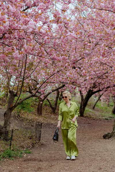 Çiçek açan pembe kiraz çiçeklerinin önünde, yeşil saten elbiseli şık sarışın bir kadın zarafet ve zarafet yayar, baharın ve doğanın güzelliğini büyüleyici bir şekilde yakalar.