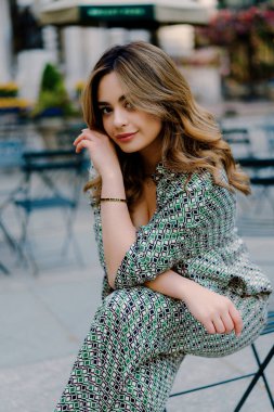 Şık desenli takım elbiseli şık bir kadın şehir parkındaki bir sandalyede oturuyor, elleri saçında, arka planda büyük bir bina var, şık ve şık bir görünüm sergiliyor. New York