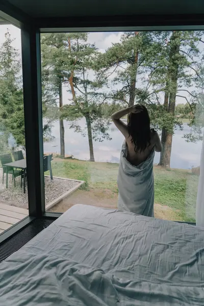 Yatağın yanında duran bir kadın yorganla kaplı bir pencereden dışarı bakıyor. Göl ve ormanın panoramik manzarası. Kırsal yazlık. Arkadan tanınmayan bir kız..