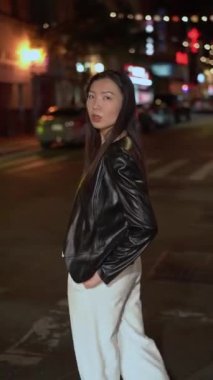 Kendine güvenen bir manken moda bir deri elbiseyle poz veriyor, gece canlı bir şehir arka planına karşı.