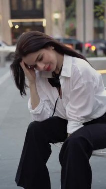 Beyaz gömlekli ve siyah pantolonlu kendine güvenen bir kadın, bir şehir sokağında oturuyor, gülümsüyor ve düşünceli görünüyor. Profesyonel ve kentin yoğun geçmişinde dengeli görünüyor.