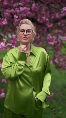 Gözlüklü zarif bir kadın çiçek açan ağaçların altında poz verir. Yeşil saten bir bluz giyer. Moda, yaşam tarzı ve canlı bahar görüntülerine ihtiyaç duyan doğa projeleri için mükemmel.