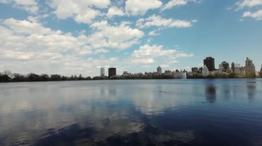 Sakin bir kentsel göl, parlak mavi gökyüzünün altında tüylü bulutları ve çarpıcı bir modern şehrin ufuk çizgisini yansıtır. New York Merkez Parkı.