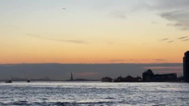 Sakin suların üzerinde gün batımının nefes kesici ve huzurlu görüntüsü uzaktaki bir şehrin ufuk çizgisini güzel bir şekilde gösteriyor. New York 'ta Özgürlük Heykeli.