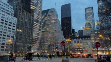 Aydınlatılmış gökdelenler ve hareketli bir meydan içeren New York şehrinin göz kamaştırıcı bir akşam manzarası..