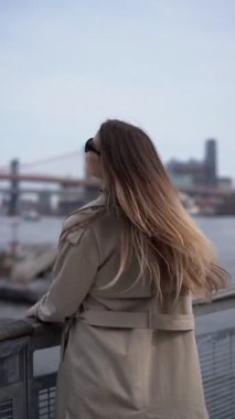 Şık bir trençkot giymiş bir kadın sakin bir şehir ortamında parıldayan suyun kenarındaki uzak bir şehir köprüsüne bakıyor. Trençkot giyen şık bir kadın suyun kenarında durmuş güzel şehir köprüsüne bakıyor.