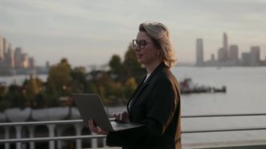 Profesyonel bir kadın şehir silueti arka planı olan, üretkenliği ve şehir yaşam tarzını somutlaştıran bir dizüstü bilgisayar kullanır..
