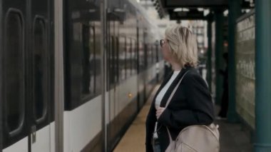 Şık, kendine güvenen bir kadın şık bir tren istasyonunda zarif bir şekilde bekler, şık kıyafetlerini gösterir ve poz verir.