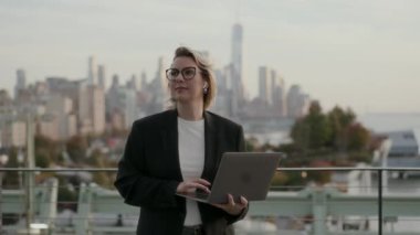 Profesyonel bir kadın şehir silueti arka planı olan, üretkenliği ve şehir yaşam tarzını somutlaştıran bir dizüstü bilgisayar kullanır..