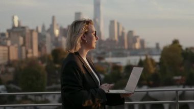 Profesyonel bir kadın şehir silueti arka planı olan, üretkenliği ve şehir yaşam tarzını somutlaştıran bir dizüstü bilgisayar kullanır..
