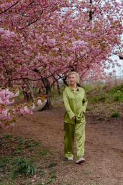 Çiçek açan pembe kiraz çiçeklerinin önünde, yeşil saten elbiseli şık sarışın bir kadın zarafet ve zarafet yayar, baharın ve doğanın güzelliğini büyüleyici bir şekilde yakalar.