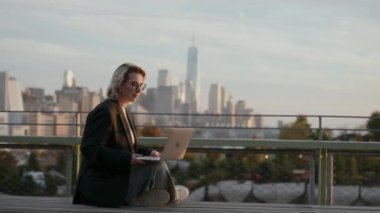 Profesyonel bir kadın şehir silueti arka planı olan, üretkenliği ve şehir yaşam tarzını somutlaştıran bir dizüstü bilgisayar kullanır..