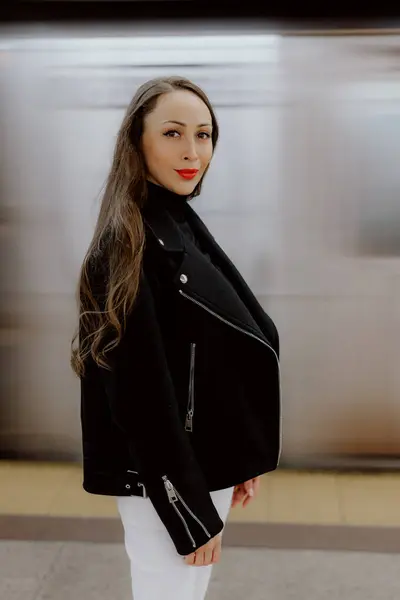 Siyah ceket giyen şık ve kendine güvenen bir kadın kalabalık bir metro istasyonunda zarafetle poz veriyor.