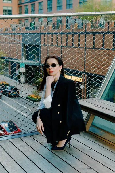 Siyah ceket ve güneş gözlüğü takmış şık bir kadın şehrin arka planına karşı şehir balkonunda kendine güvenen bir duruş sergiliyor şık ve sofistike bir görünüm sergiliyor.