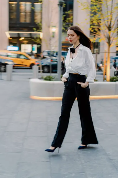 Beyaz bluz ve siyah pantolon giyen başarılı bir iş kadını, hareketli şehirde güven ve profesyonellik yayar. Kurumsal kıyafetinin başarısını ve zarafetini simgeler.