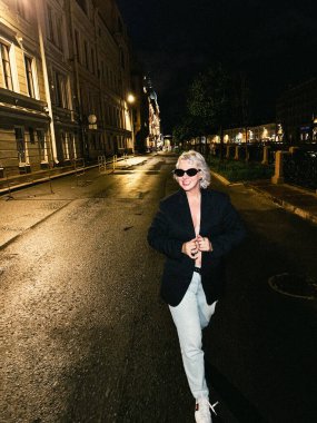 Şık bir birey, hareketli bir gecede loş bir şehir caddesinde rahatça dolaşır. Şehirde Şık ve Şık Bir Gece Yürüyüşü